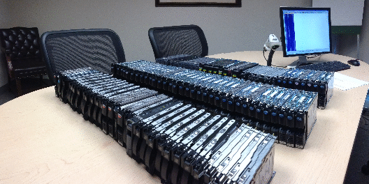 Photograph of a conference table covered with rows of hard drives. A computer, scanner, and chairs are also visible in the background.